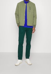 Uomo che indossa un giubbotto imbottito oliva, una camicia blu reale, pantaloni verde scuro e sneaker bianche, in piedi davanti a uno sfondo bianco.