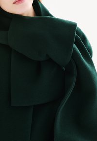 Tumma vihreä villahuivi, jossa on suuri, pehmeä solmusi yksityiskohta, teksturoitu pinta ja laskostettu kangas, joka luo virtaavan ilmeen.