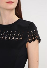 Robe noire avec un col rond, des manches courtes dotées de motifs circulaires et ovales découpés, et une texture de tissu douce.