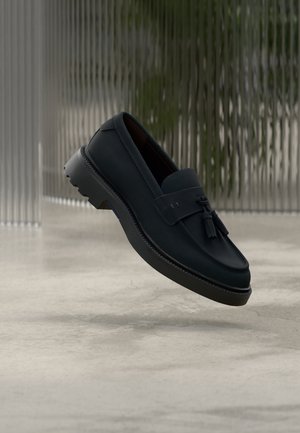 Mörkblå läderloafers med en slät yta, försedd med en dekorativ tofs och en robust svart gummisula, upphöjda från marken.