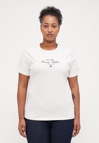 T-shirt en coton blanc avec un col rond, des manches courtes et un texte de logo imprimé violet. Porté avec un jean en denim bleu foncé.