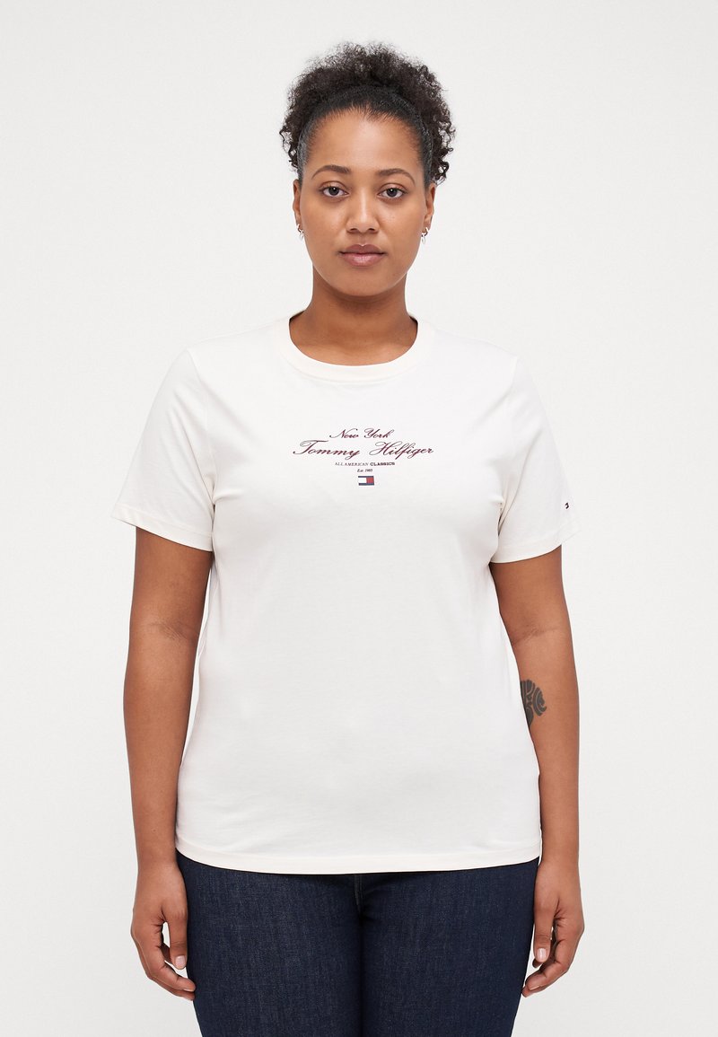 T-shirt en coton blanc avec un col rond, des manches courtes et un texte de logo imprimé violet. Porté avec un jean en denim bleu foncé.
