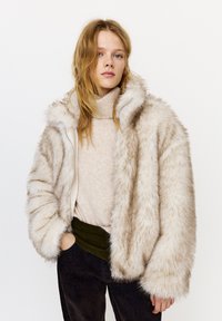 Fiatal nő laza szőke hajjal bézs garbót, túlméretezett világos szőrme kabátot és sötét nadrágot visel, fehér háttér előtt állva.