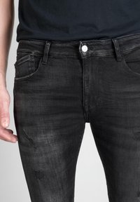 Jeans en denim noir avec une coupe slim, présentant des décolorations subtiles, des patches usés et un design classique à cinq poches. Fermeture par bouton à la taille.