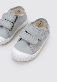 Chaussures en tissu gris avec deux bandes velcro et semelles en caoutchouc blanches. Elles présentent un dessus texturé et un renfort à l'avant pour la durabilité.