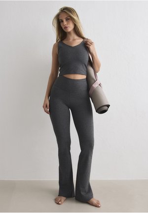 Kvinne i grå ermeløs crop top og vide leggings som holder en rullet yogamatte over skulderen, stående barbent mot en ensfarget vegg.