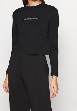 Vrouw draagt een zwart cropped top met lange mouwen met de tekst "CALVIN KLEIN JEANS" en losse zwarte broek, staand tegen een effen achtergrond.