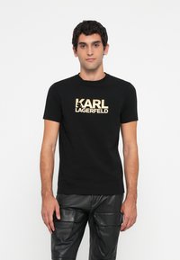 Melna kokvilnas t-kreklu ar lielu zelta "KARL LAGERFELD" logo priekšā. Īsas piedurknes un apaļš kakla izgriezums, kombinēts ar melnām ādas biksēm.