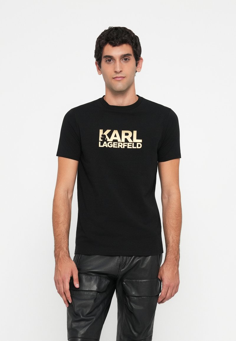 Melna kokvilnas t-kreklu ar lielu zelta "KARL LAGERFELD" logo priekšā. Īsas piedurknes un apaļš kakla izgriezums, kombinēts ar melnām ādas biksēm.