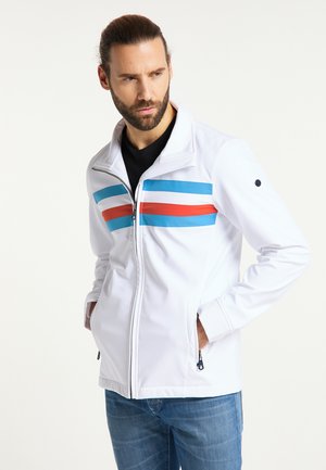 Mann mit Bart, der eine weiße Jacke mit roten und blauen Streifen trägt, Hände in den Taschen, schaut seitlich vor einfachem Hintergrund.
