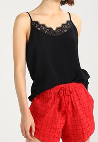 Kvinna iförd en svart linne med spetsdetaljer och röda texturerade shorts med dragsnöre, stående mot en enkel ljus bakgrund.