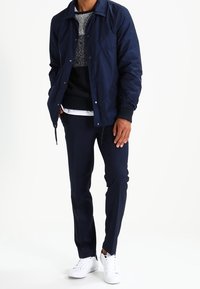 Marineblau leichtgewichtige Jacke mit Druckknöpfen, strukturierter schwarzer Pullover, weißes Unterhemd, dunkle Hose und weiße Sneakers.