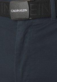 Pantaloni blu navy con delicate righe diagonali, dotati di una cintura nera texturizzata con una fibbia rettangolare incisa con "CALVIN KLEIN."
