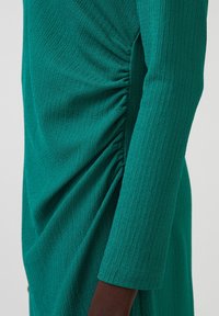 Abito teal con maniche lunghe, caratterizzato da un tessuto testurizzato e dettagli arricciati lungo il lato, che mette in risalto una silhouette aderente.