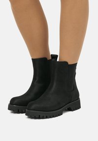Botas negras de ante hasta el tobillo con suelas gruesas y con relieve, llevadas sobre piernas desnudas contra un fondo blanco.