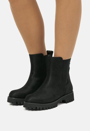 Botas negras de ante hasta el tobillo con suelas gruesas y con relieve, llevadas sobre piernas desnudas contra un fondo blanco.
