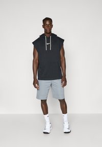Felpa senza maniche nera con tasca frontale, cordini color crema, abbinata a pantaloni corti grigio chiaro e sneaker bianche. Logo Nike sulla felpa.