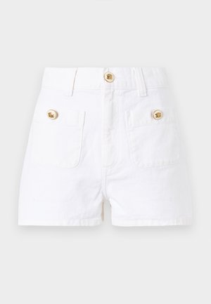 Γυναίκα που φοράει μπλε ριγέ πουκάμισο χωρίς μανίκια με δέσιμο μπροστά, λευκές σορτς, καφέ δερμάτινα παπούτσια, κρατώντας μια μικρή σκούρα καφέ τσάντα.