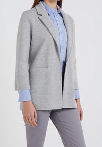 Ljust grå kavaj med slagkrage, tillverkad av mjukt tyg. Två framfickor och långa ärmar med blå och vita randiga manschetter.