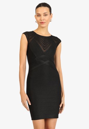 Frau trägt ein eng anliegendes schwarzes ärmelloses Kleid mit transparenten Zickzack-Mustereinsätzen an Brust und Schultern, stehend vor weißem Hintergrund.