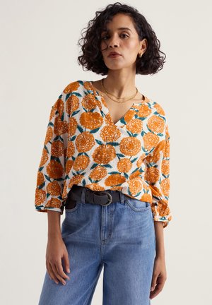 Rövid göndör hajú nő fehér blúzzal, narancssárga virágmintával, a derekánál magasított kék farmerbe tűrve, fekete övvel.