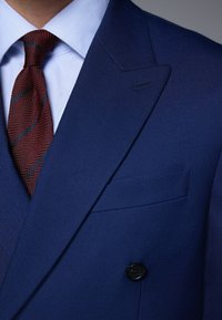 Granatowa teksturowana marynarka z klapami w szpic i zapięciem na jeden guzik, zestawiona z czerwonym krawatem w jodełkę na jasnoniebieskiej koszuli w paski.