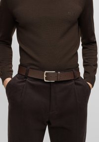 Ceinture en cuir marron avec une finition texturée, dotée d'une boucle rectangulaire en argent. Portée sur un pantalon marron foncé et un pull marron ajusté.