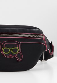 Černá látková taška s růžovým lemováním a grafickým designem, který zobrazuje obličej se zelenými slunečními brýlemi. Obsahuje stříbrný zip a nastavitelné ucho.