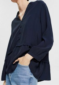 Blouse bleu marine avec col en V, boutonnée sur le devant et ourlet à volants. Manches longues avec poignets froncés et tissu léger et lisse.