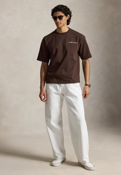 Άνδρας που φοράει σκούρο καφέ μπλουζάκι Polo Ralph Lauren, λευκό παντελόνι, γυαλιά ηλίου, ρολόι και λευκά παπούτσια, στέκεται σε απλό φόντο.