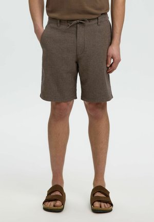 Pantalones cortos marrones rectos con bolsillos delanteros usados por una persona con una mano en el bolsillo y sandalias marrones en los pies.