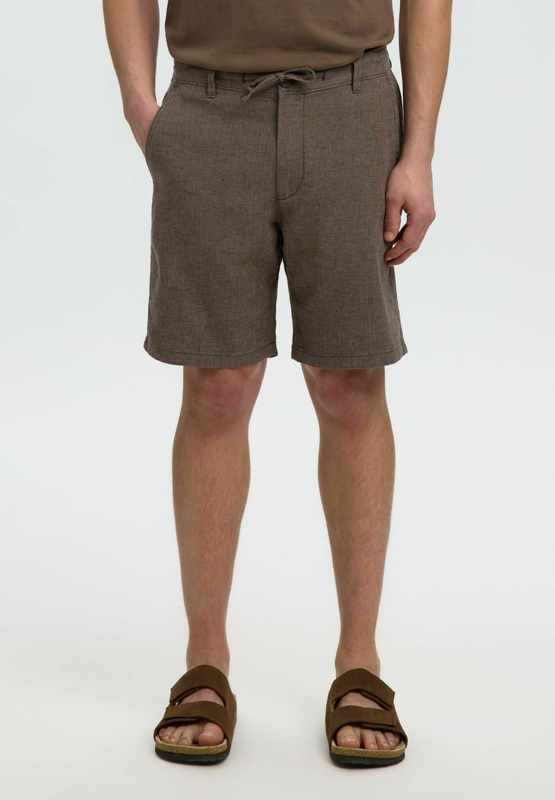 Shorts marron droits avec poches avant portés par une personne avec une main dans la poche et des sandales marron aux pieds.