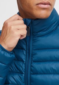 Blauer Puffermantel mit hohem Kragen und horizontalen Nähten. Mit Reißverschluss schließende Vorderseite, glatte Textur und glänzende Oberfläche.