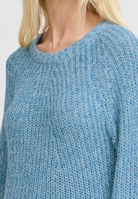 Gros plan d'une personne portant un pull en tricot bleu et gris à motifs avec un col rond côtelé.