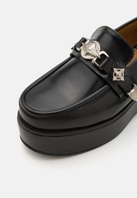 Pantofii din piele neagră, cu talpă platformă, ce prezintă elemente decorative din metal argintiu și cusături complicate de-a lungul perimetrului și al curelei.