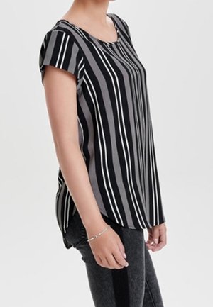 Femme portant un chemisier à manches courtes rayé verticalement noir et gris et un jean foncé, debout de profil devant un fond uni.