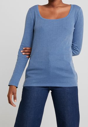 Kvinde iført en lyseblå, ribbet bluse med lange ærmer og firkantet halsudskæring samt mørkeblå vidde jeans mod en lys baggrund.