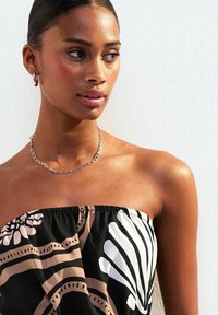 Robe sans bretelles avec une base noire, ornée de motifs floraux audacieux en blanc et beige. Collier en chaîne dorée et teint de peau uniforme.