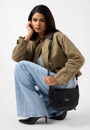 Schwarze Leder-Schultertasche mit strukturierter Oberfläche, goldenem Logo-Akzent und verstellbarem Riemen. Model trägt braune Jacke mit Kragen und Schlagjeans.