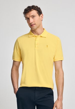 Gele polo gemaakt van zacht materiaal. Heeft een klassieke kraag, twee knopen en een klein logo op de borst. Gemaakt in combinatie met donkere broek.