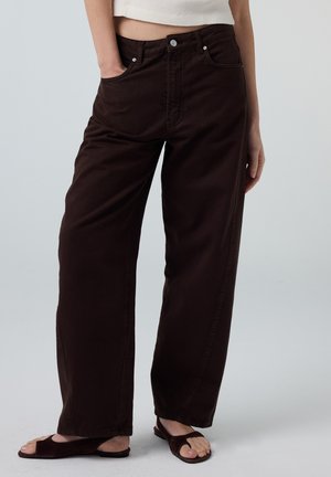 Vrouw die donkerbruine, hoog getailleerde, wijd uitlopende broek draagt met een witte cropped top en donkerbruine open-toe sandalen, één hand in de zak.