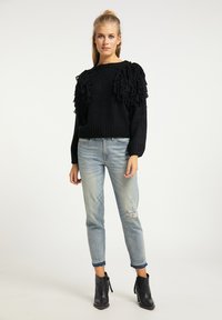 Pulover tricotat gros, de culoare neagră, cu accente franjurate pe umeri, asortat cu blugi deschiși, deteriorați, de culoare albastră și cizme negre până la gleznă.