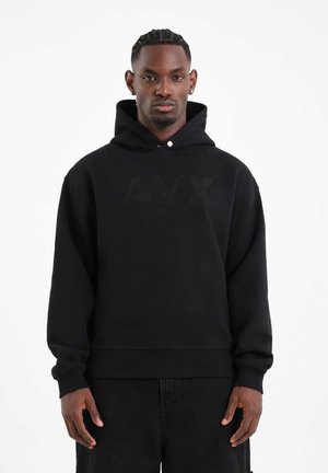 Uomo con capelli intrecciati che indossa una felpa nera con cappuccio AVRX e pantaloni neri, in piedi davanti a uno sfondo bianco uniforme.