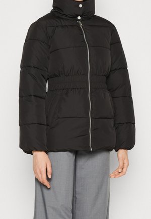 Doudoune noire avec col montant, fermeture éclair devant, taille élastique et manches longues, portée avec un pantalon gris clair.