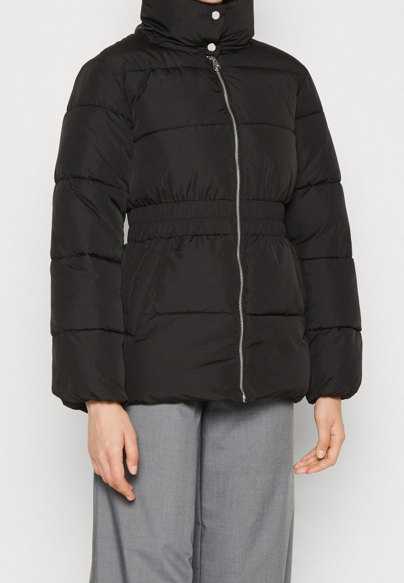 Doudoune noire avec col montant, fermeture éclair devant, taille élastique et manches longues, portée avec un pantalon gris clair.