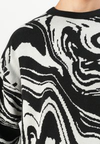 Zwart-witte gebreide trui met een abstract spiralenmarmerpatroon, ronde hals, gedragen door een persoon die van schouder tot kin is afgebeeld.
