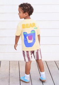 Yellow T-shirt with blue bear graphic and "BEAR HUS" text; striped shorts in brown and white, blue sneakers, wooden deck background.