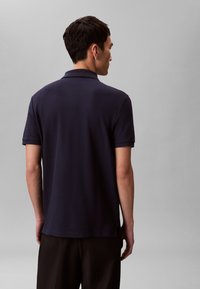 Férfi, aki sötétkék, rövid ujjú galléros pólót és fekete nadrágot visel, háttal áll egy egyszínű szürke háttér előtt.