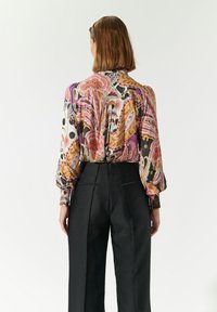 Blusa colorata a fantasia con maniche lunghe, pantaloni neri. La blusa presenta motivi floreali e astratti in tonalità di rosa, viola e arancione.