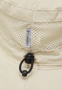 Beige aurinkohattu, jossa on verkko-osa, säädettävä leukahihna ja Columbia-logo. Kangas on sileä, ja lippavarren ympärillä on ompeluakkentteja.